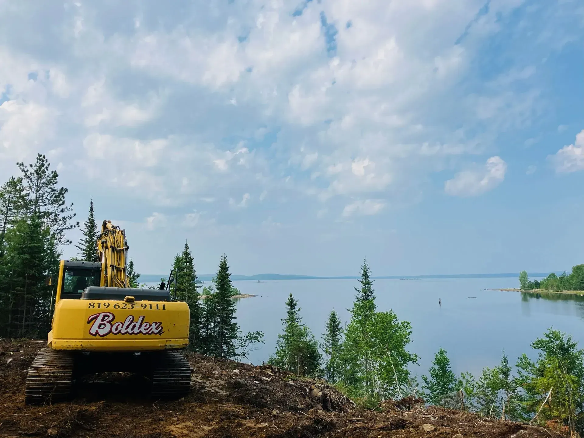 Une pelleteuse jaune sur une colline surplombant un lac et des arbres sous un ciel nuageux.