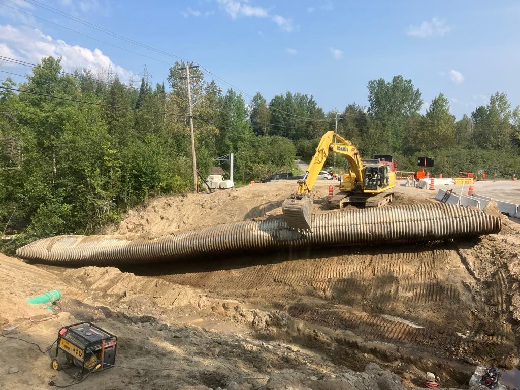 Une pelle mécanique jaune travaille sur un grand ponceau en tôle ondulée lors d'une excavation. En plein jour, à l'extérieur.