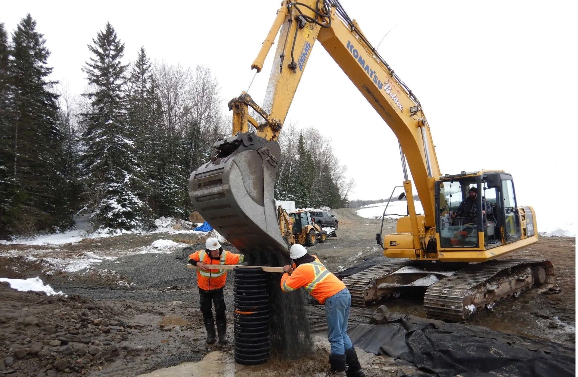 Des ouvriers du bâtiment installent des tuyaux de drainage à l'aide d'une excavatrice dans une zone boueuse, avec une forêt enneigée en arrière-plan.