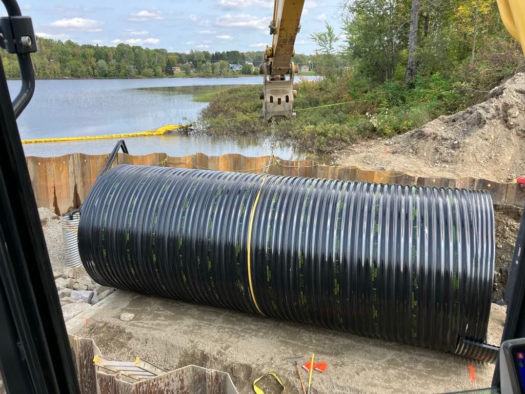 Chantier : une excavatrice place un gros tuyau ondulé dans une zone partiellement creusée près d'un lac.