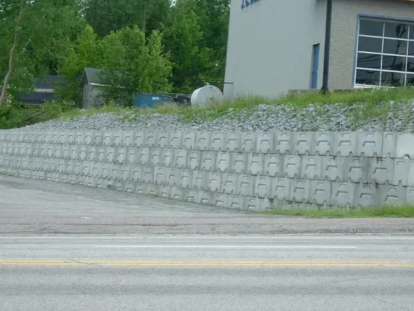 Mur de soutènement en blocs de béton gris le long d'une route. Un bâtiment et des arbres se trouvent à l'arrière-plan.