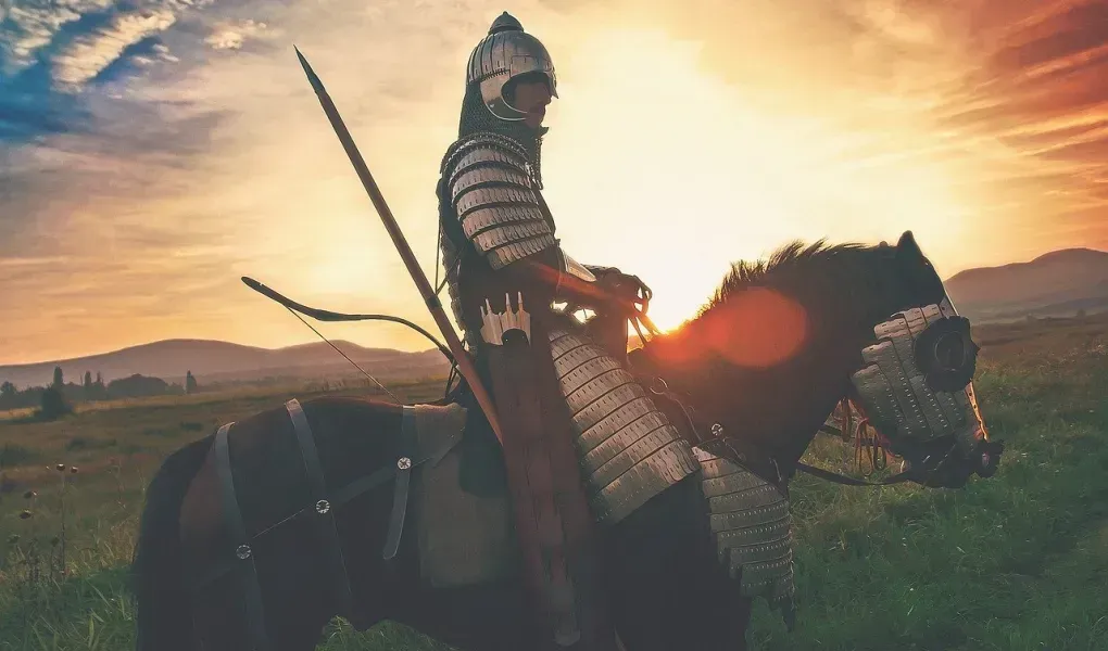 A man in armor is riding a horse in a field at sunset.