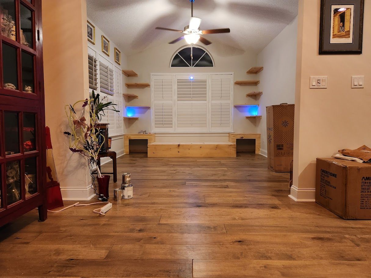 Warm living room with hardwood floors, white shutters, ceiling fan, and small decor on tables and shelves.