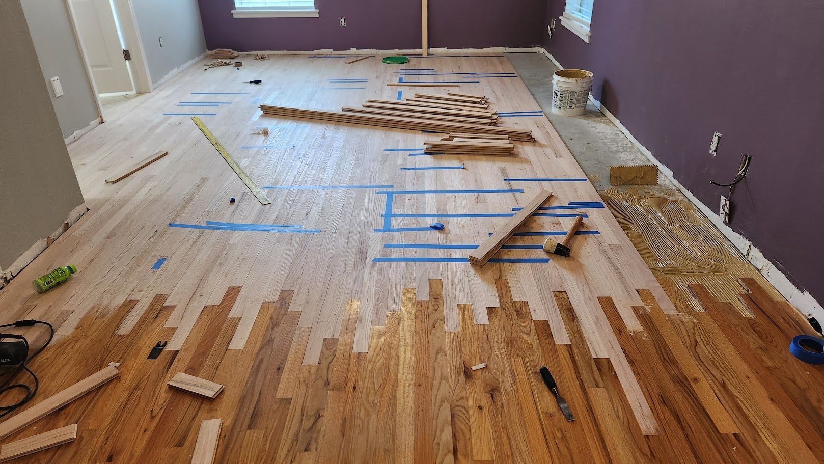 Partially installed wood floor with scattered planks and blue tape in a room with purple walls