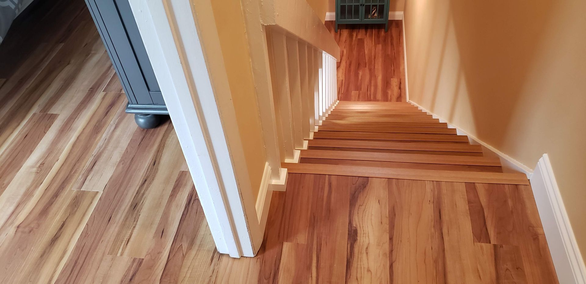 A staircase with light brown wood flooring, leading down to a landing with a dark vent cover on the wall.