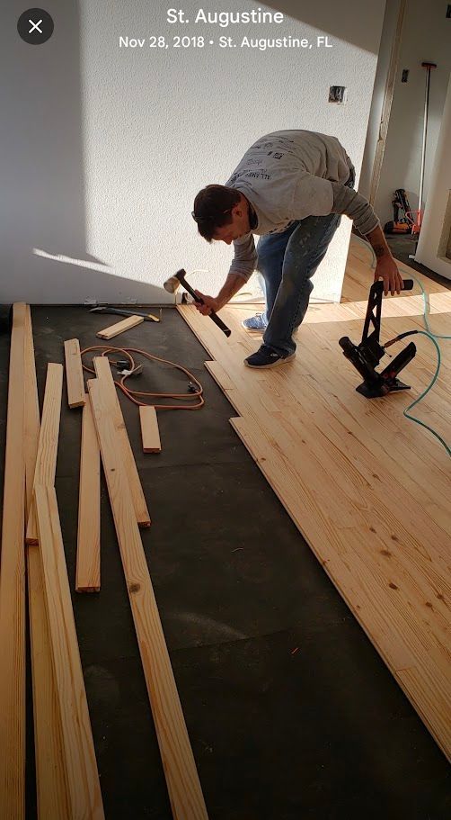 Person installing hardwood flooring in a sunlit room, with planks and tools on the floor.