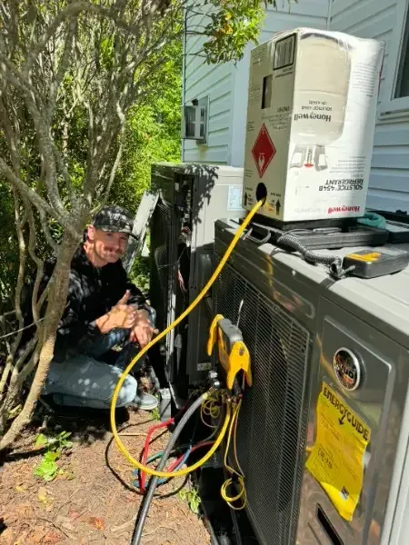 Technician working on outdoor HVAC unit beside house, with hoses and tools connected.