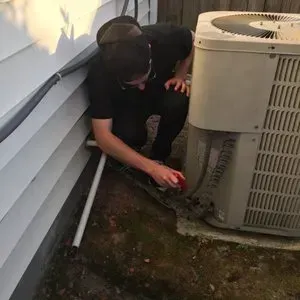 Person crouches beside an outdoor HVAC unit, inspecting a hose or cable near a house wall.
