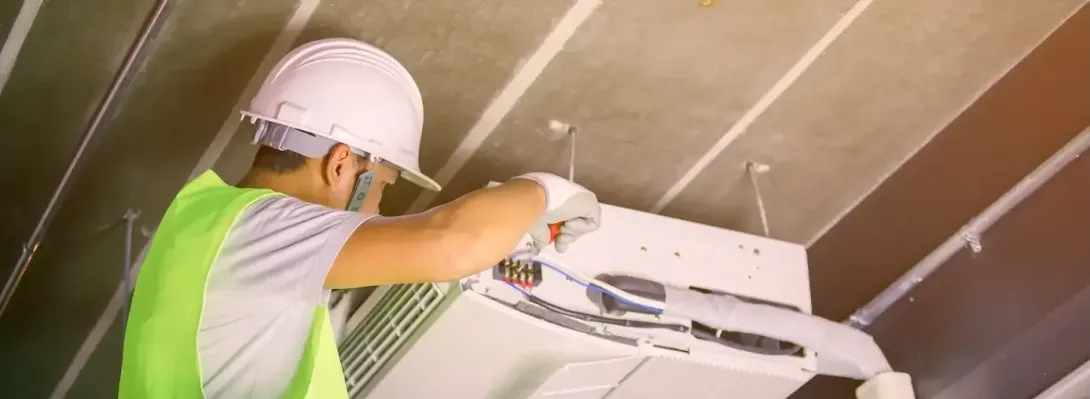 Worker in hard hat and safety vest installing a ceiling air conditioner inside a room