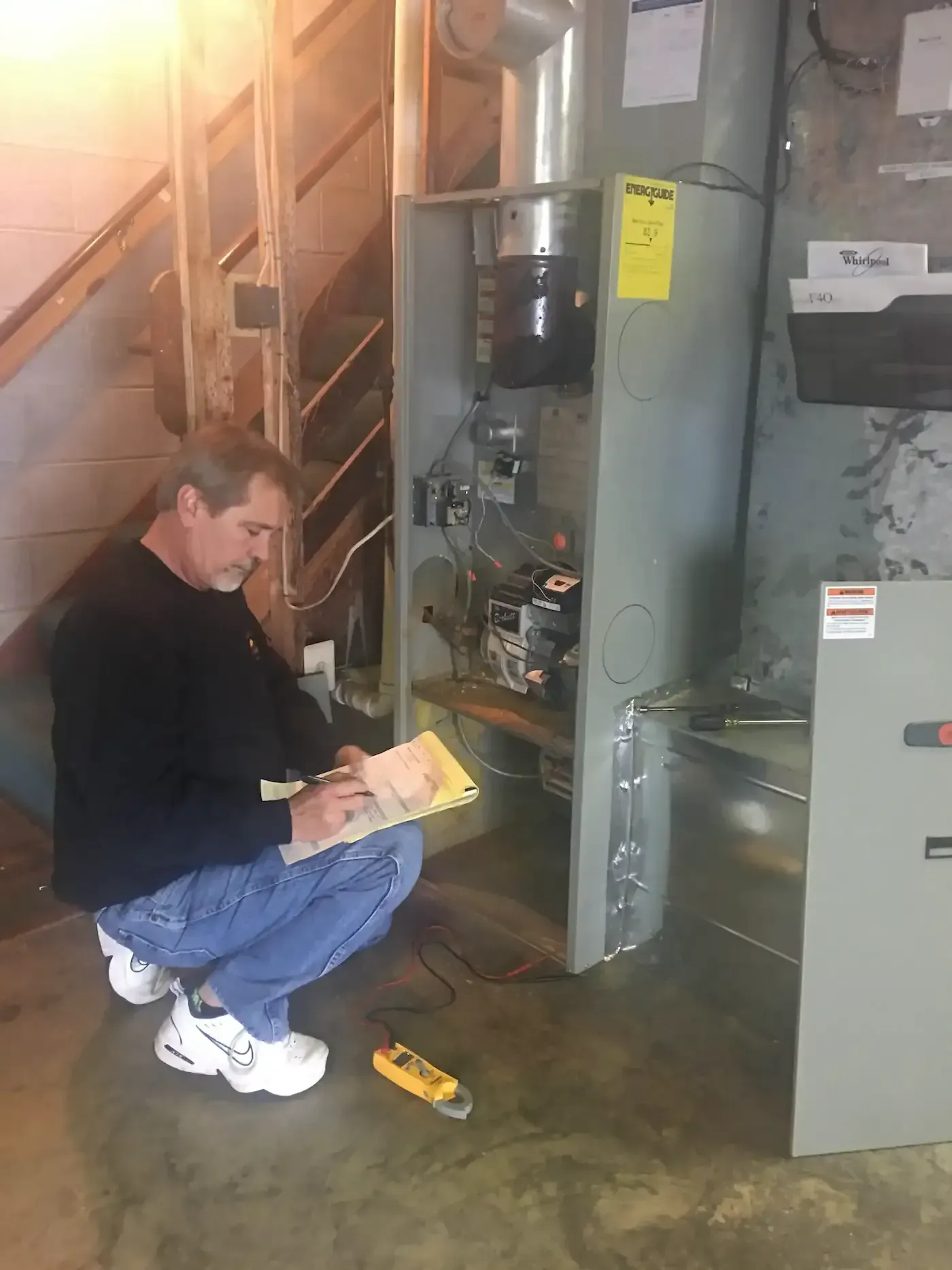 Technician crouches by an open electrical panel in a basement, inspecting wiring with tools nearby