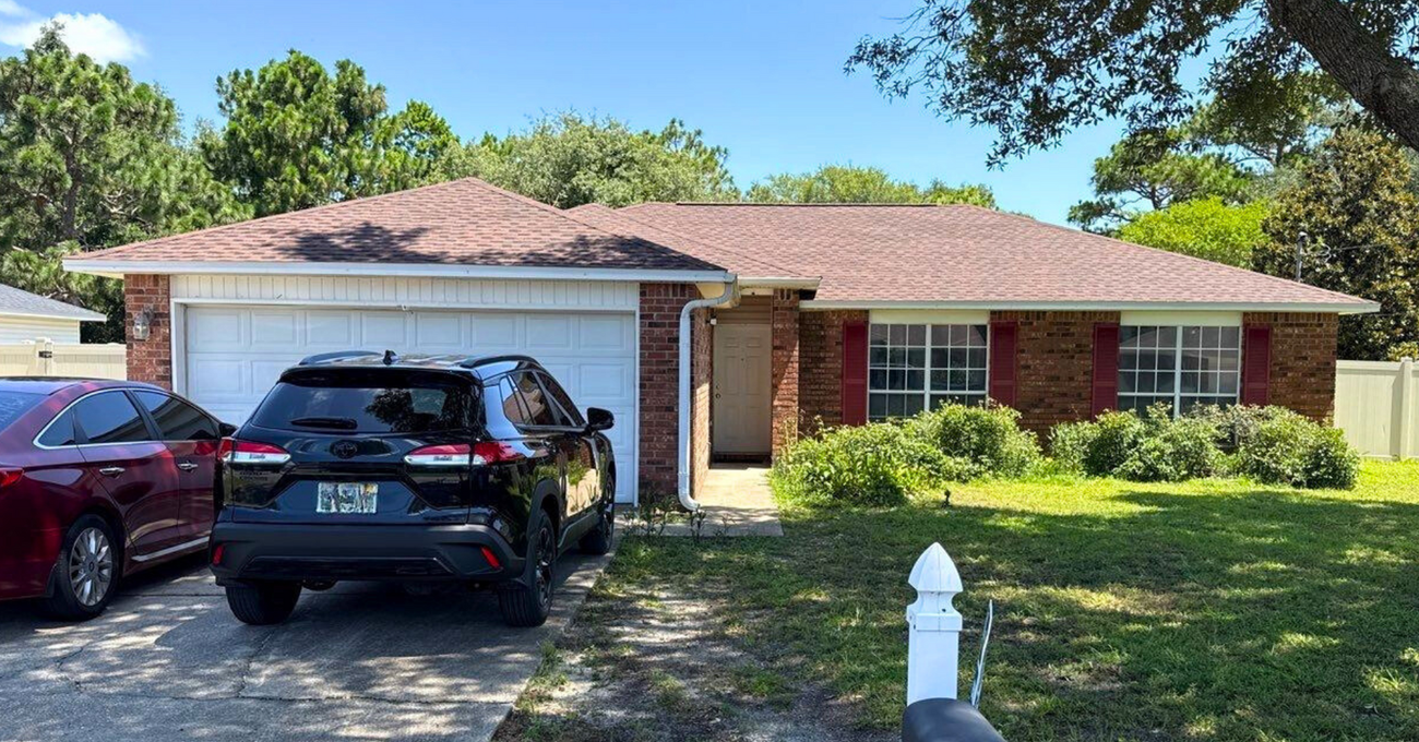 Single-story suburban house with two cars parked in the driveway, front lawn, and bright trees.