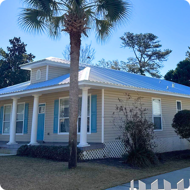 Single-story beige house with blue shutters, white porch columns, and a metal roof under palm trees