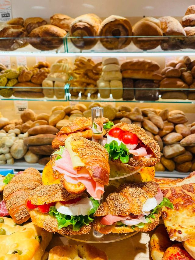 Vetrina da forno con pane assortito e un panino a strati su pane ai semi davanti