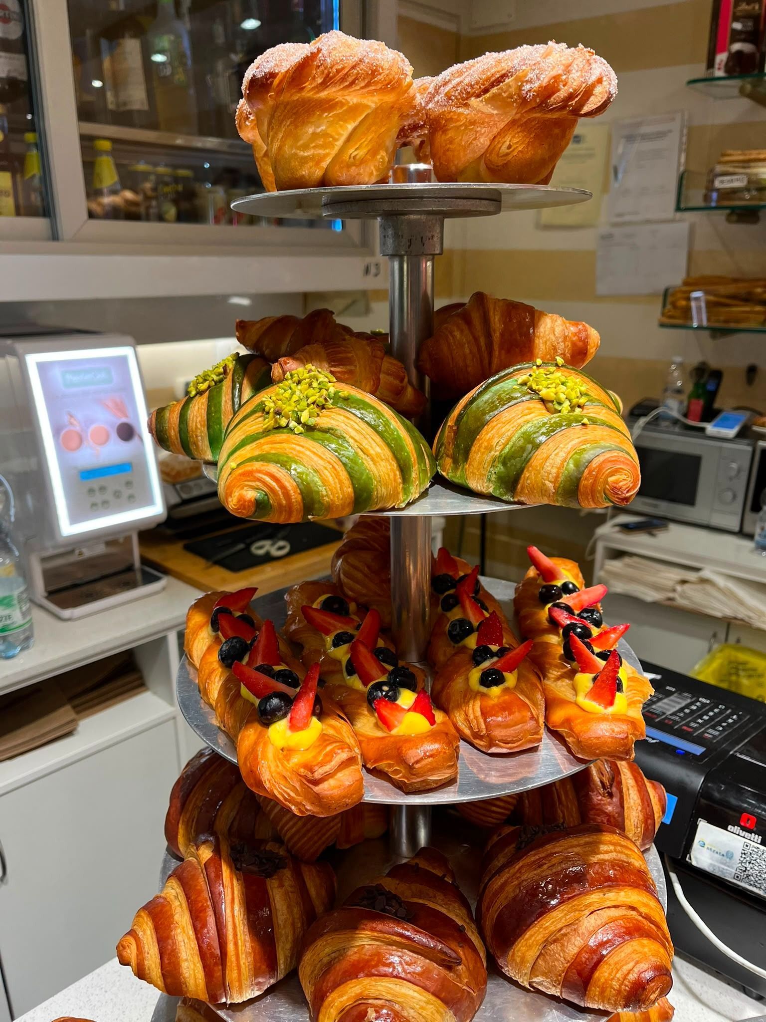 Vetrina da pasticceria con esposizione a più livelli di pasticcini e croissant colorati.