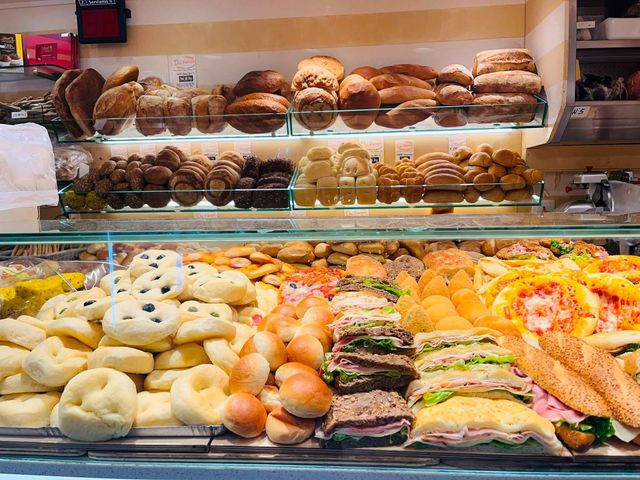 Vetrina da forno con assortimento di pane, pasticcini e panini disposti su ripiani e vassoi.
