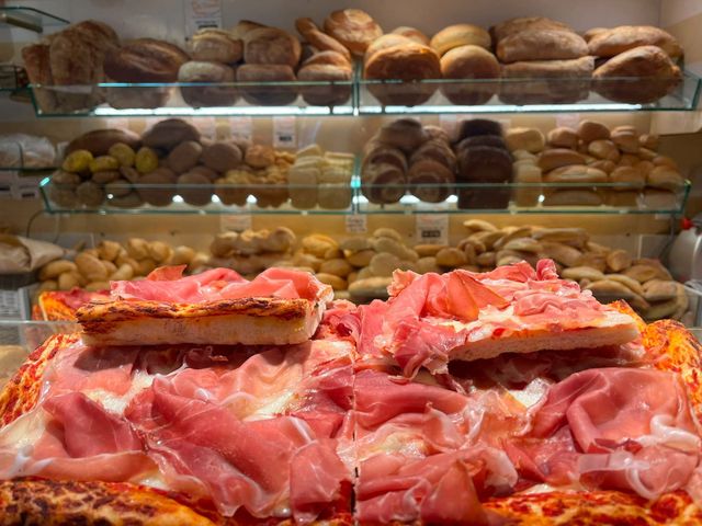 Banco espositivo con un assortimento di pane e pasticcini dietro una vetrina, con salumi affettati in primo piano.
