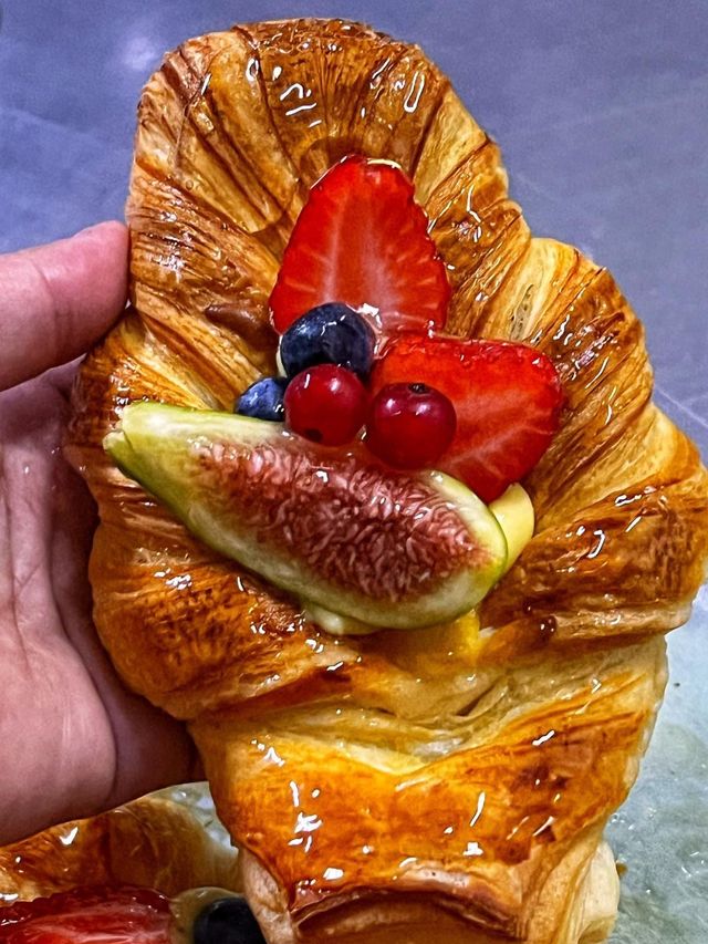 Mano che regge un croissant lucido guarnito con fragole a fette, mirtilli e una fetta di fico.