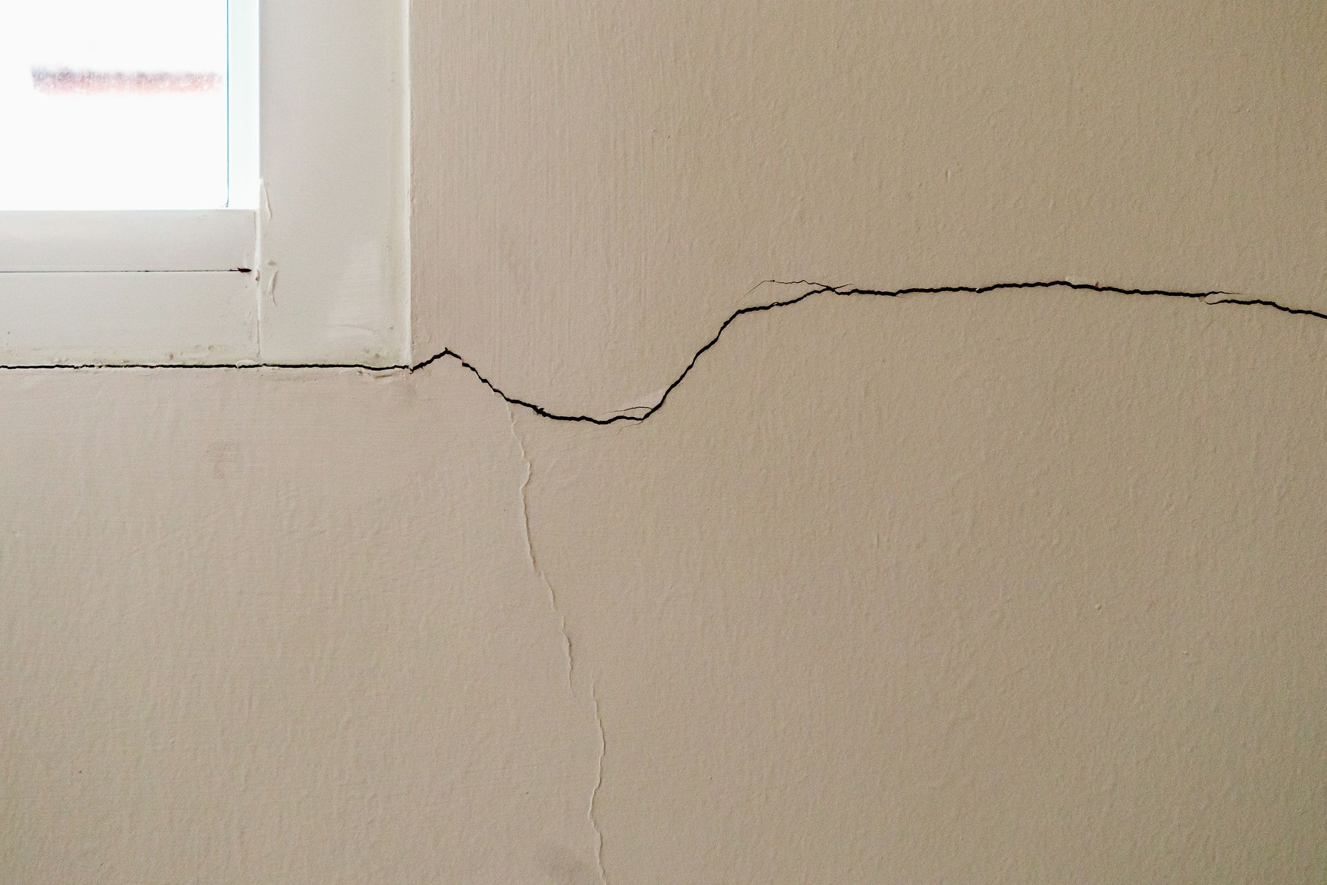Foundation Repair — Abateplus structural damage on a beige plaster wall beside a window.
