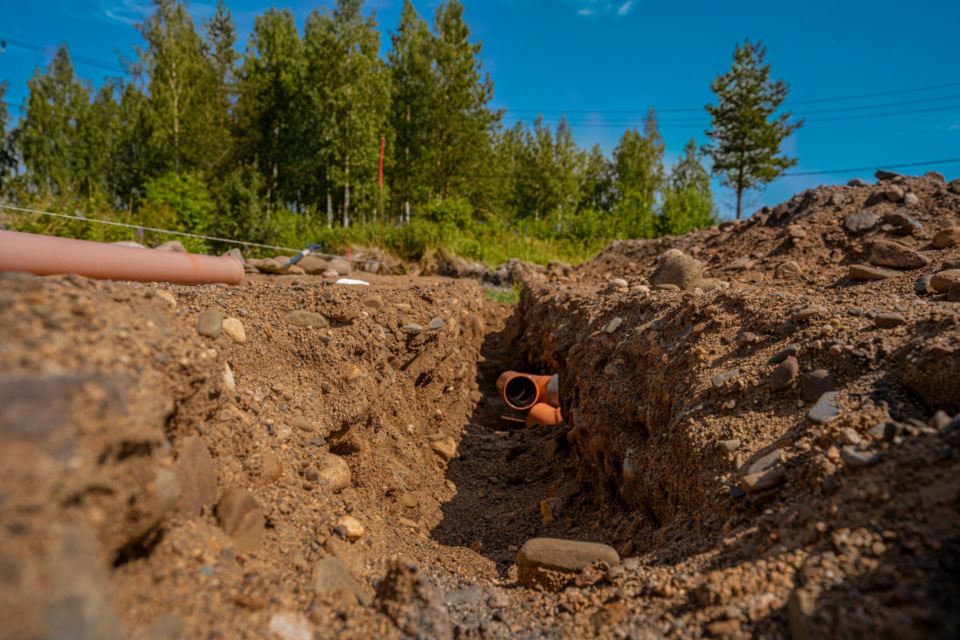 Trench with an orange pipe opening in a dirt excavation, backed by trees under a blue sky