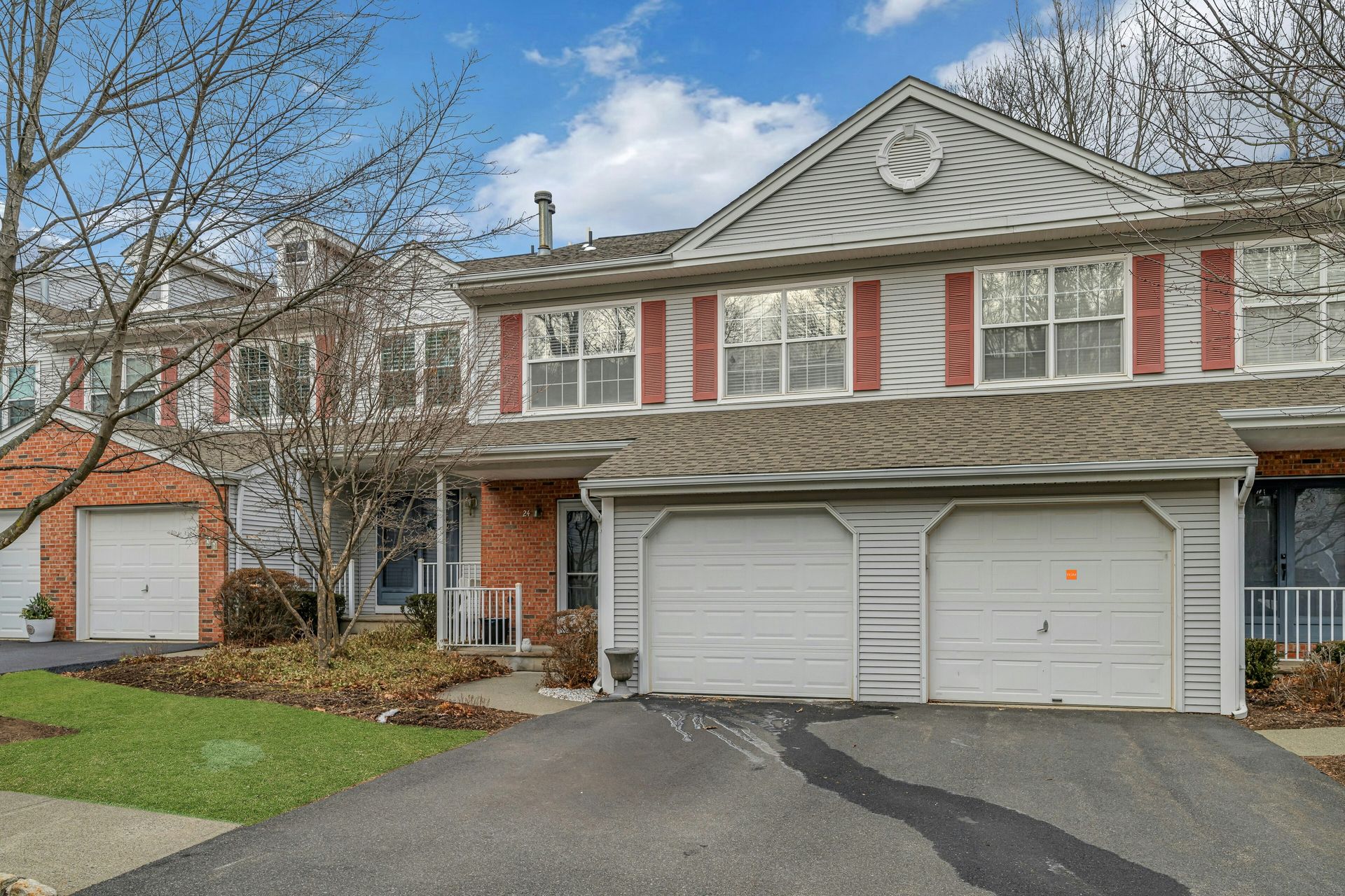 Townhouse with two-car garage, red shutters, and brick facade on a driveway with bare trees in the background.