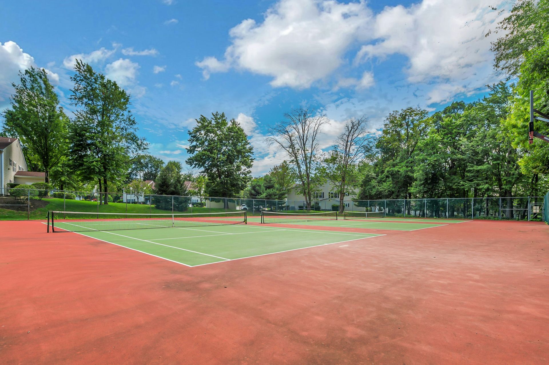 Red and green outdoor tennis court on a sunny day, trees and houses in the background.
