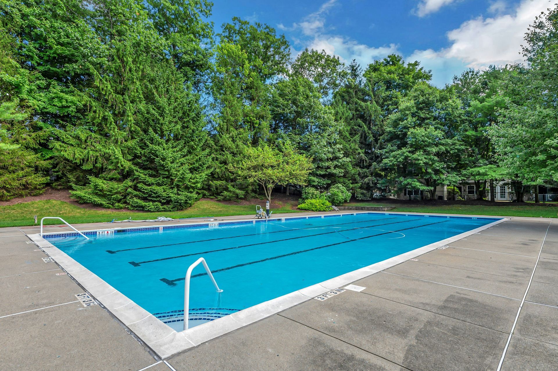 Outdoor swimming pool with blue water, surrounded by concrete and trees under a partly cloudy sky.