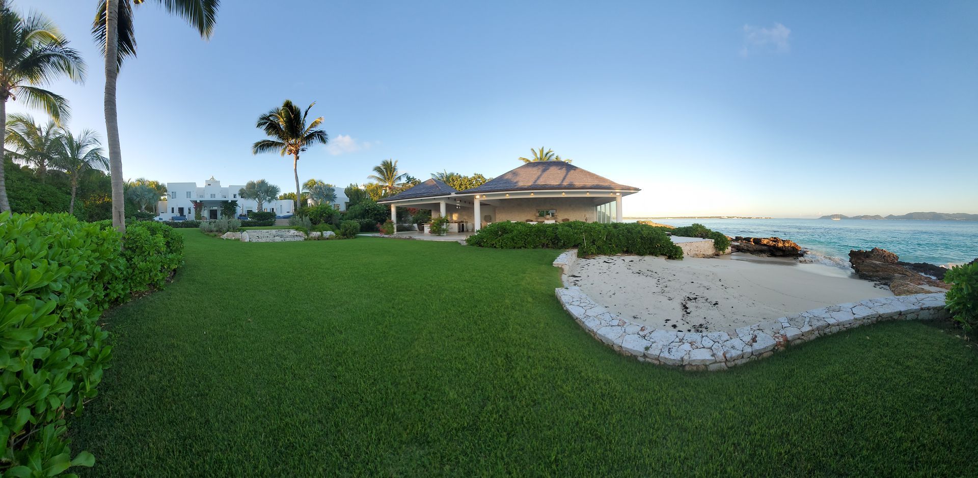 Lush green lawn with a beachfront pavilion, white house and the sea under a blue sky.