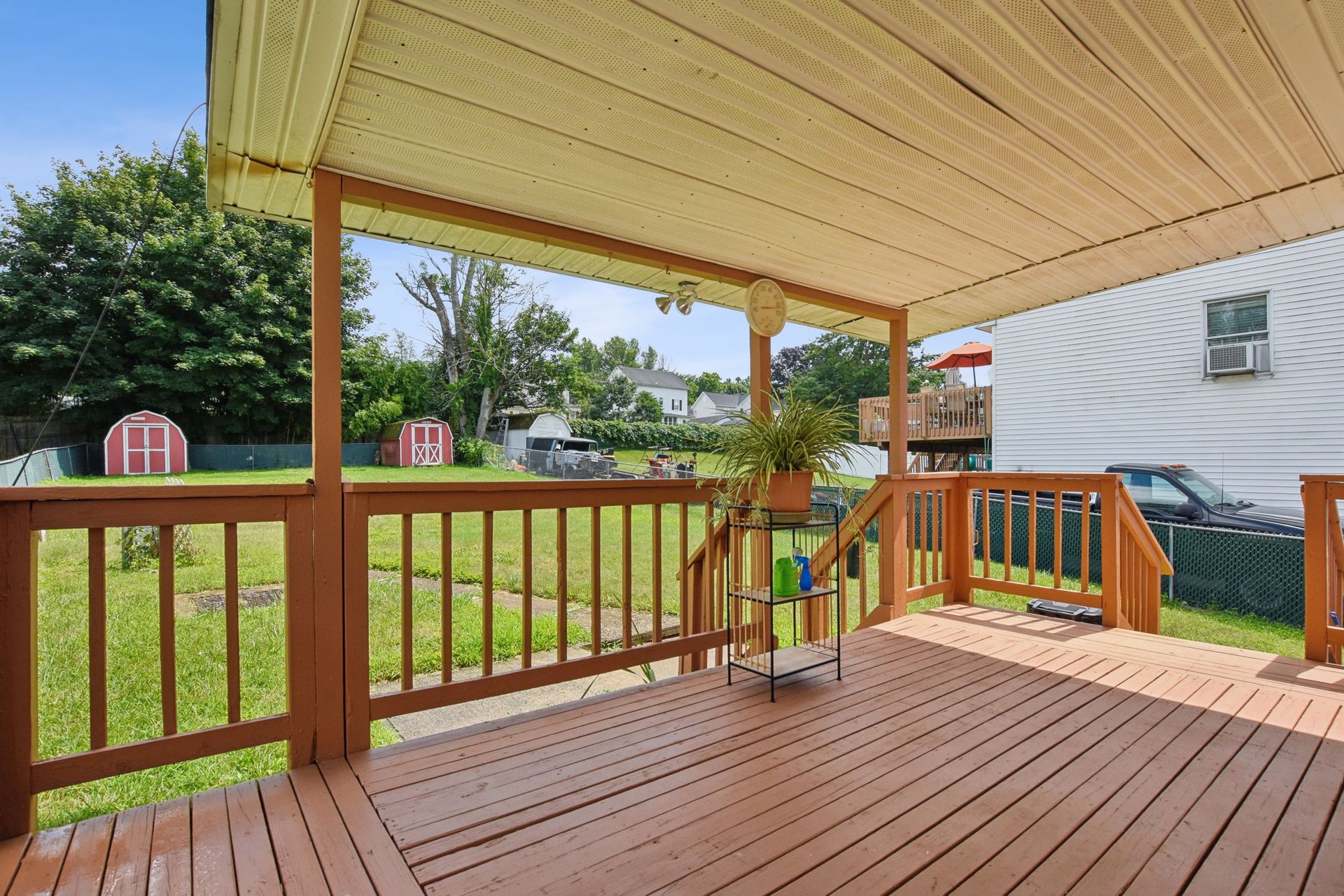 Covered wooden porch overlooking a grassy yard with sheds and houses in the background.