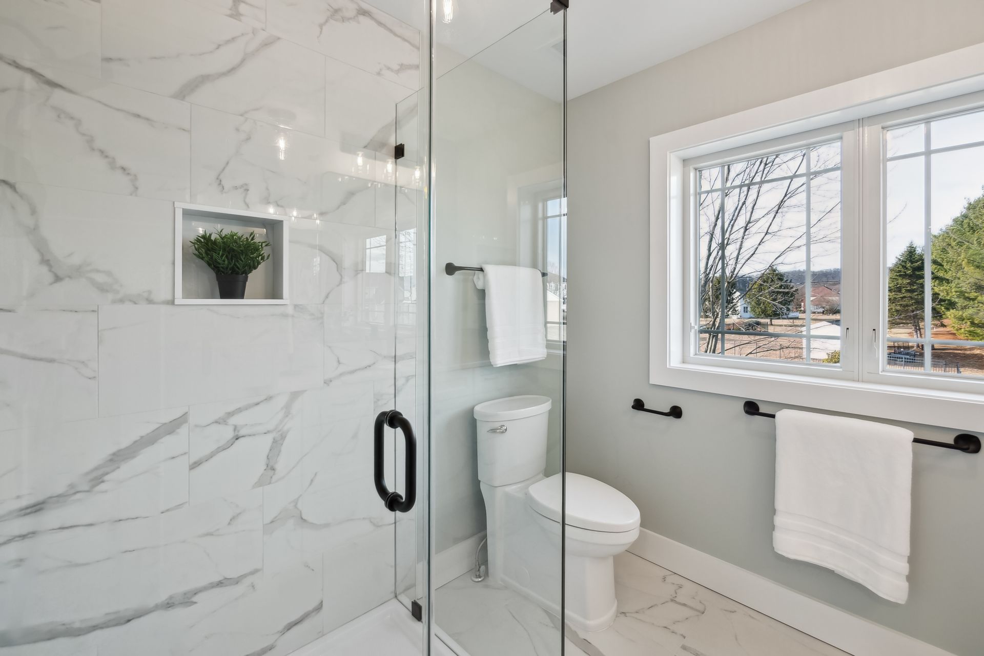Modern bathroom with marble shower, toilet, window. White towels on black bars.