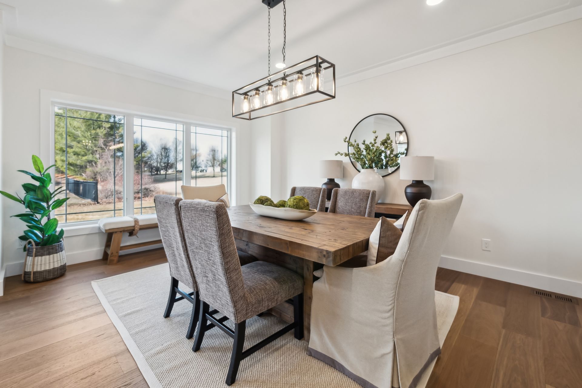 Dining room with wooden table, chairs, and rectangular light fixture. Large window, rug, and plant.