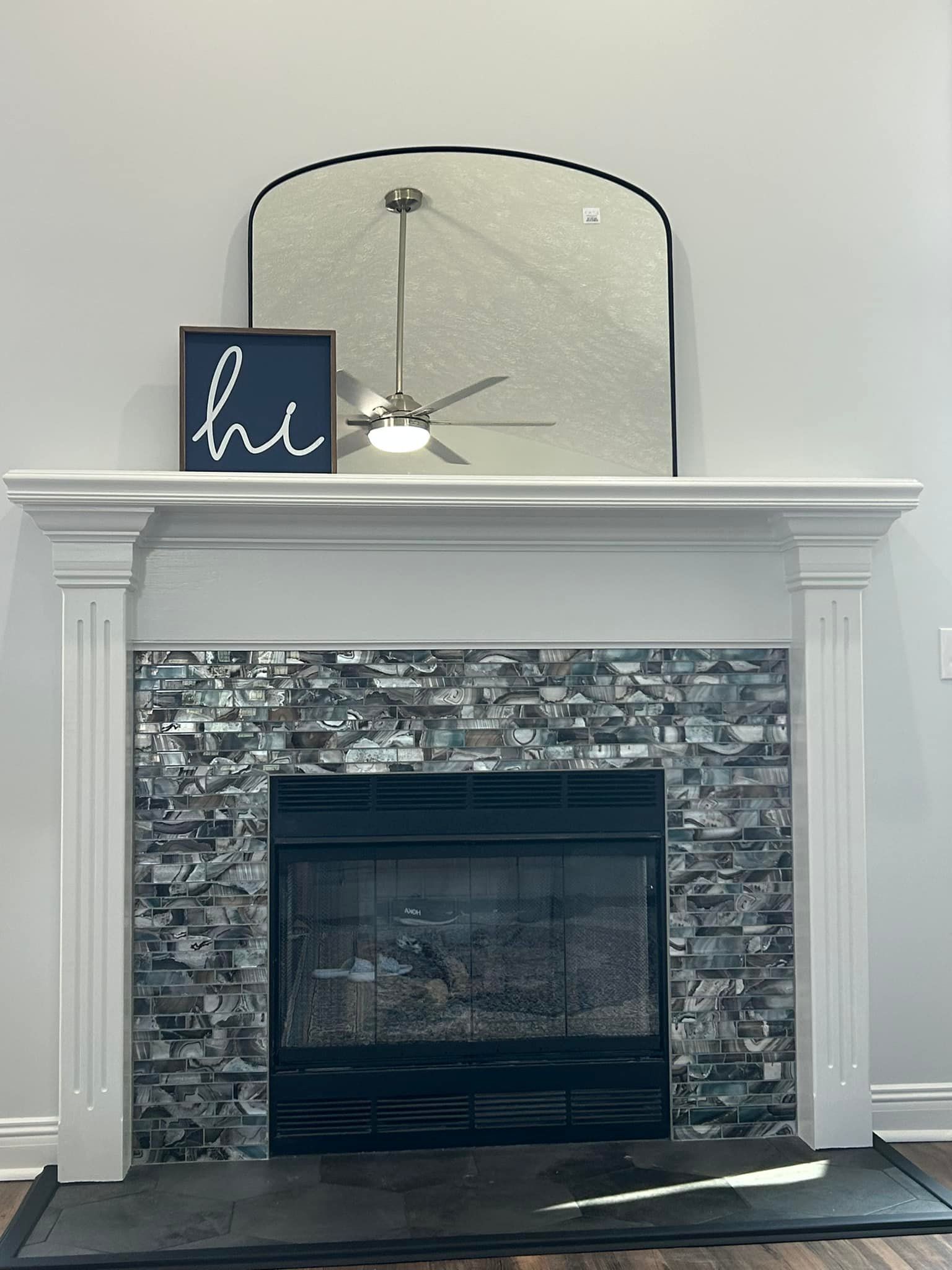 Fireplace with white mantel, gray stone surround, mirror, and ceiling fan above; small blue “hi” sign on mantel