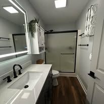 Narrow modern bathroom with white vanity, black fixtures, shower door, towel wall art, and wood-look floor.