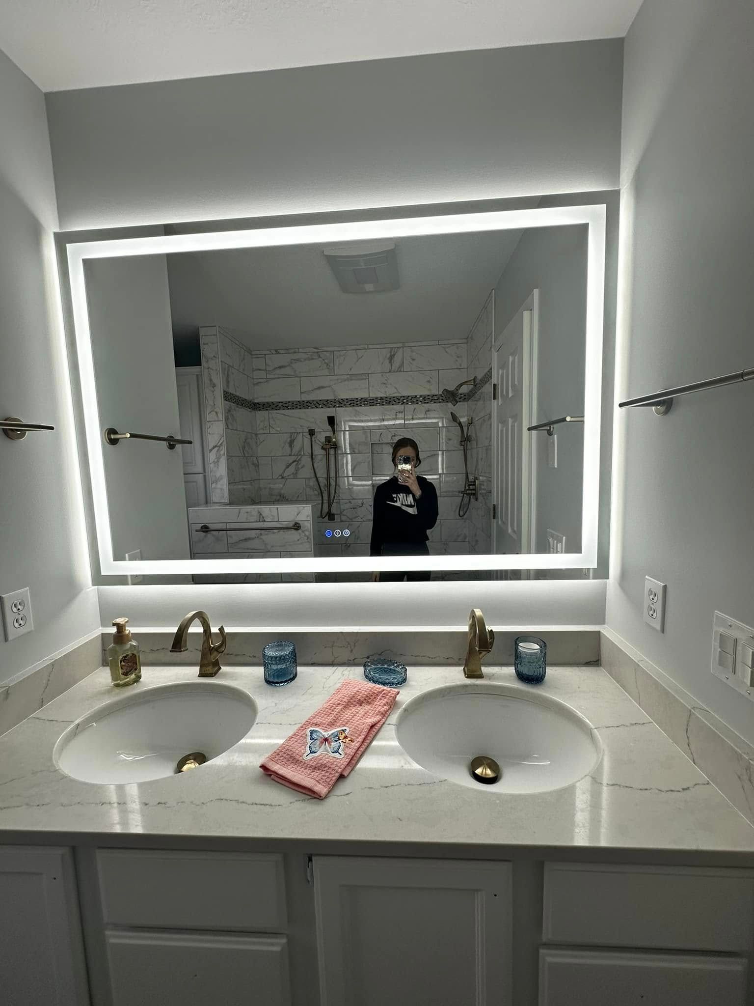 Bathroom vanity with two sinks and a lit mirror; a person is reflected standing in the background.