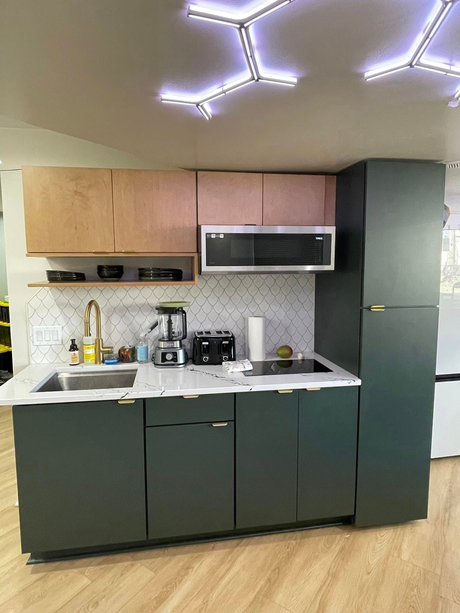 Compact modern kitchen with dark green cabinets, wood upper cabinets, white countertop, and geometric ceiling lights