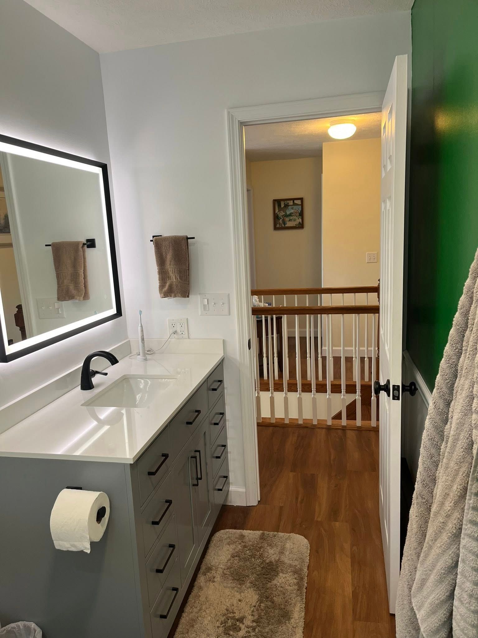 Bathroom vanity with mirror, sink, gray drawers, towel rack, and a view into a hallway with wood railing.