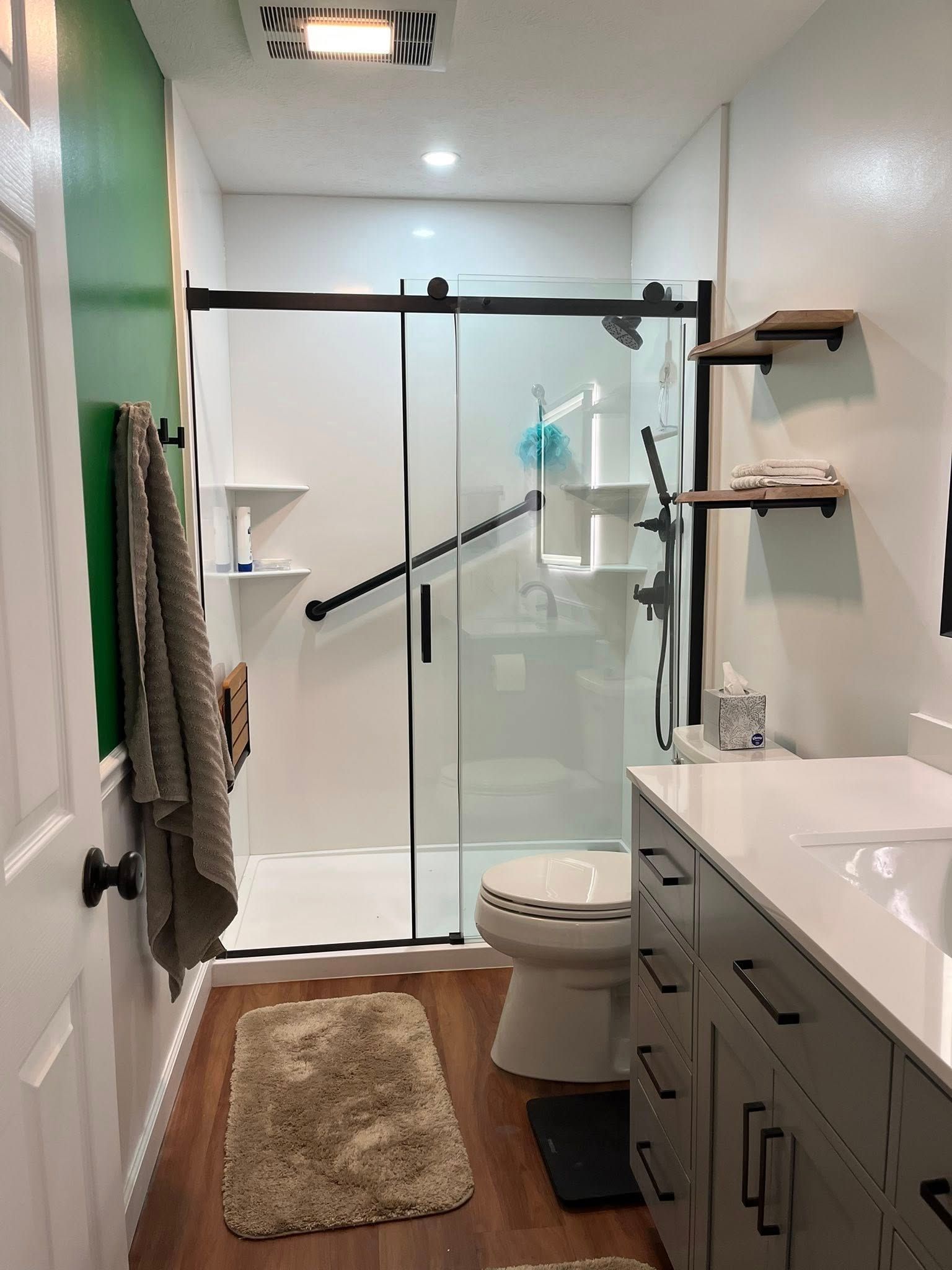 Narrow modern bathroom with glass shower, toilet, vanity, and a beige rug on a wood floor.