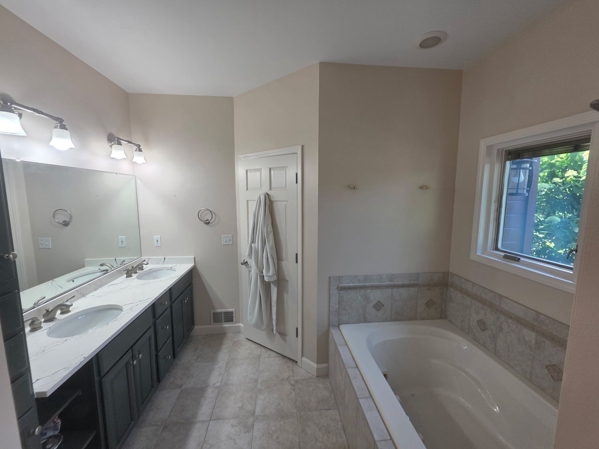 Spacious beige bathroom with double vanity, large mirror, and a soaking tub by a window.