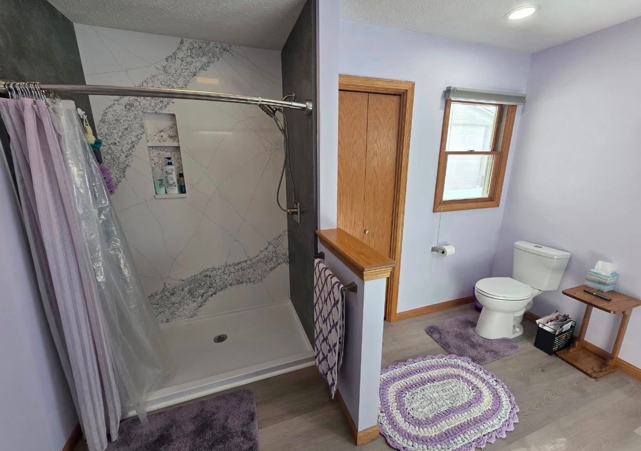 Bathroom with shower, toilet, lavender walls, purple rugs, and a small window beside a wooden door.