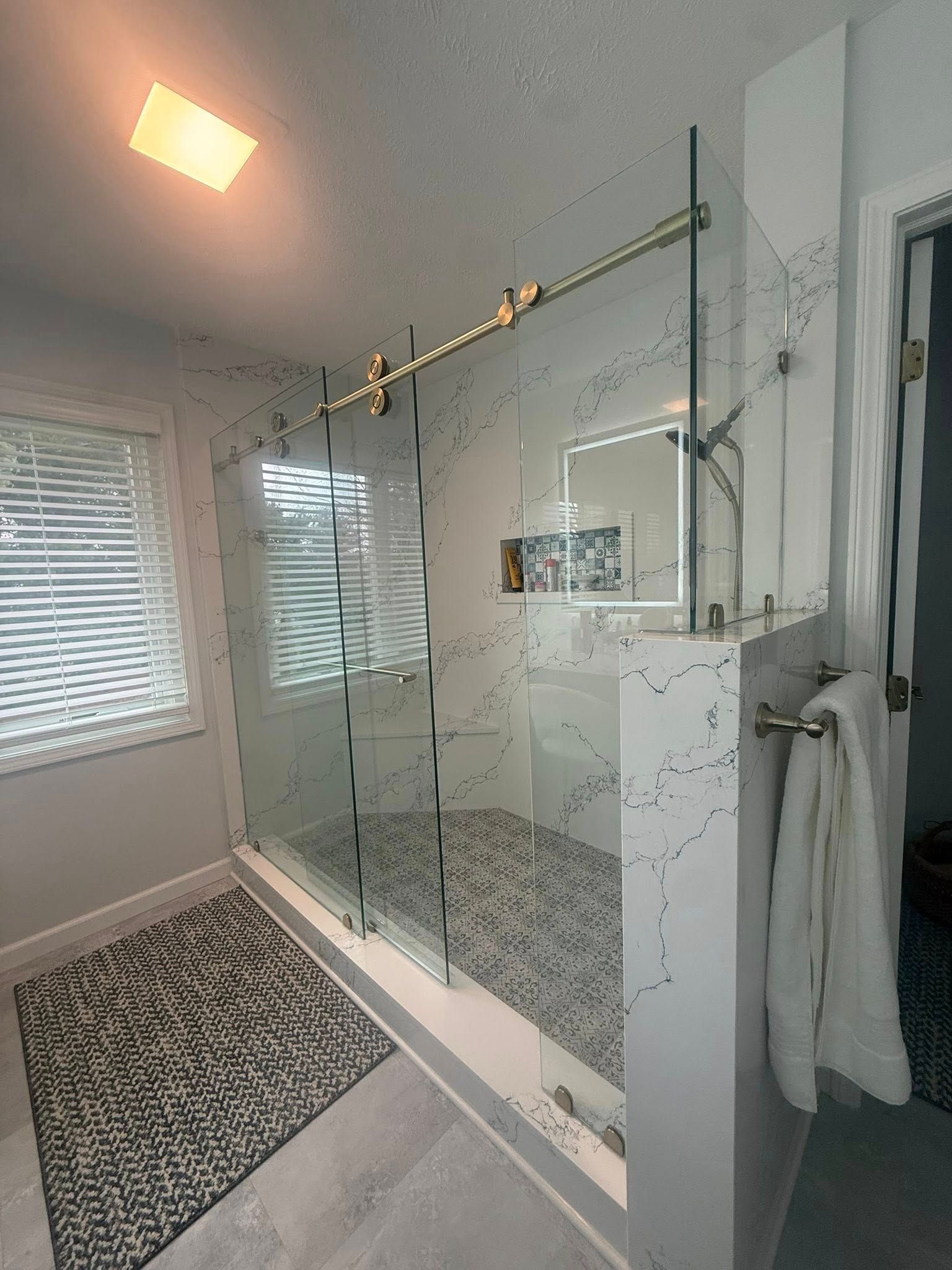 Modern bathroom with glass shower, gray tile, window blinds, and hanging towels.