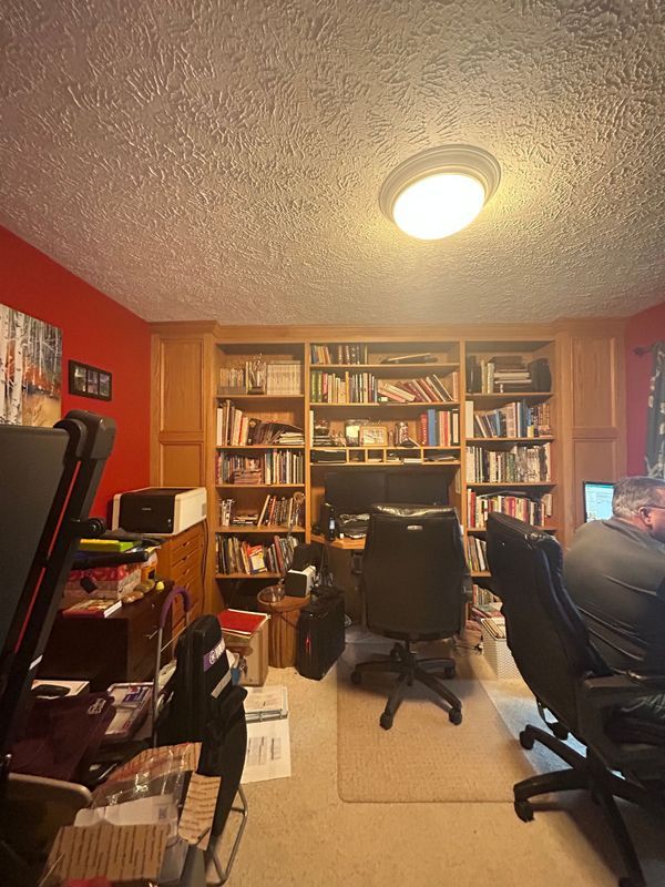 Home office with bookshelf, desk, and two black chairs on a beige rug under a ceiling light.