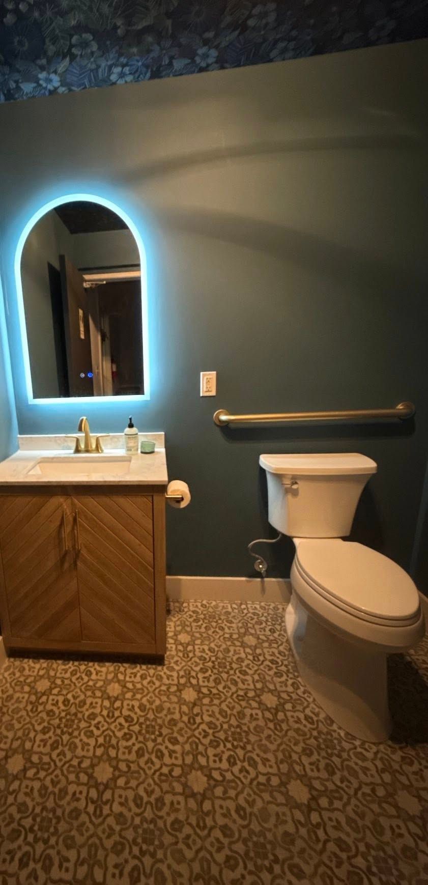 Small bathroom with a wooden vanity, arched mirror, blue wall, and toilet beside a grab bar.
