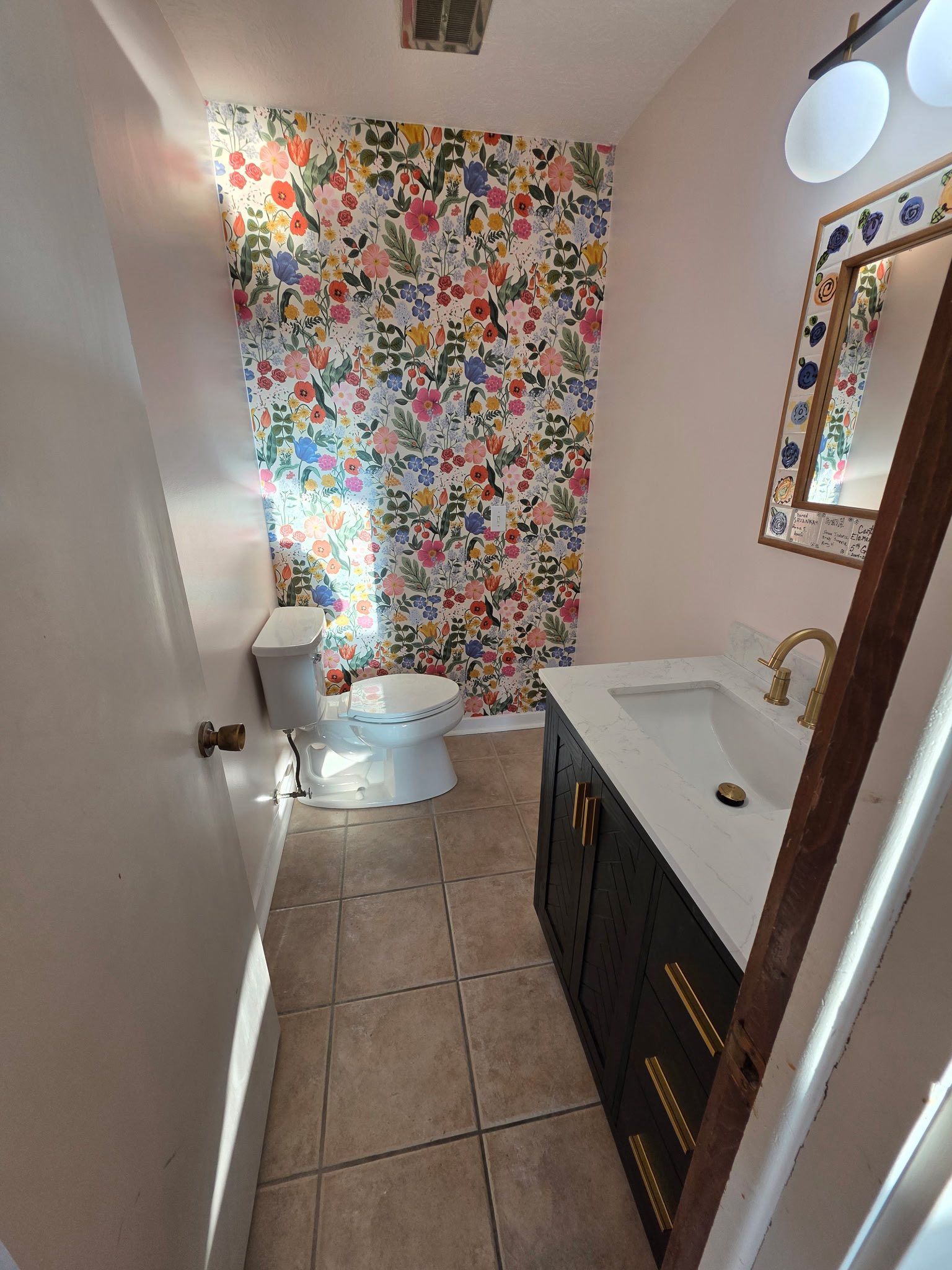 Narrow bathroom with colorful floral wallpaper, toilet, tiled floor, vanity sink, and mirror.