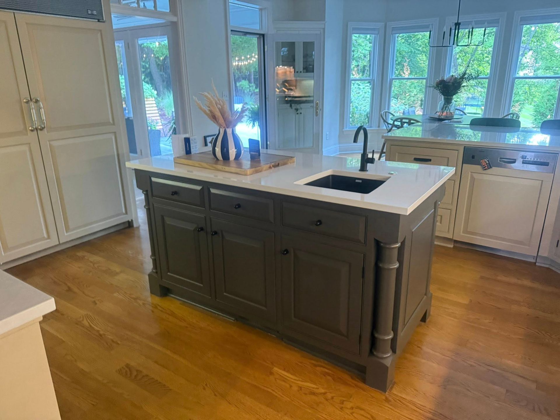 Bright kitchen with a dark island, white cabinets, wood floors, and large windows overlooking greenery