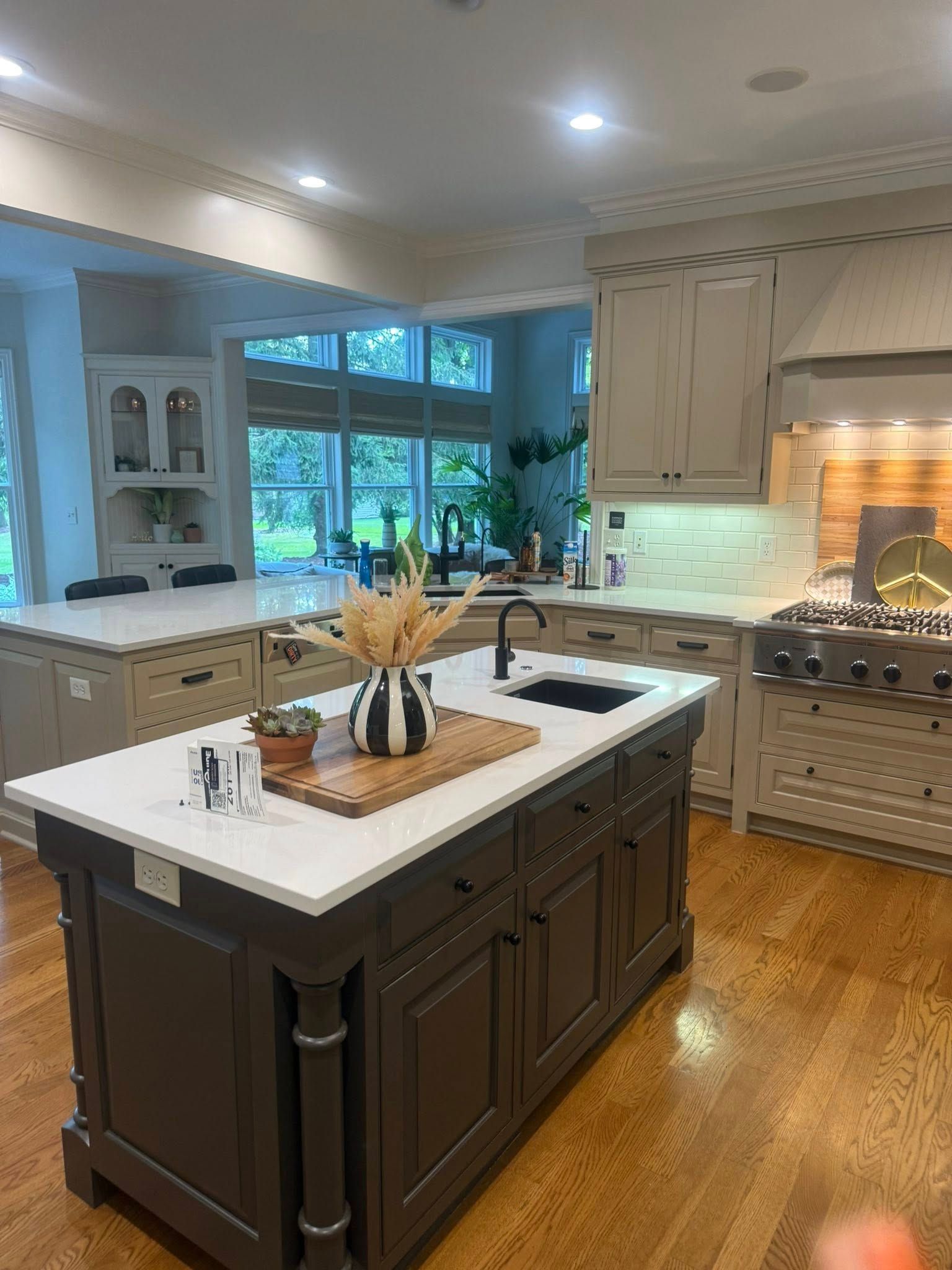 Bright modern kitchen with dark island, wood floors, large window, and stainless steel stove by a lit brick fireplace.