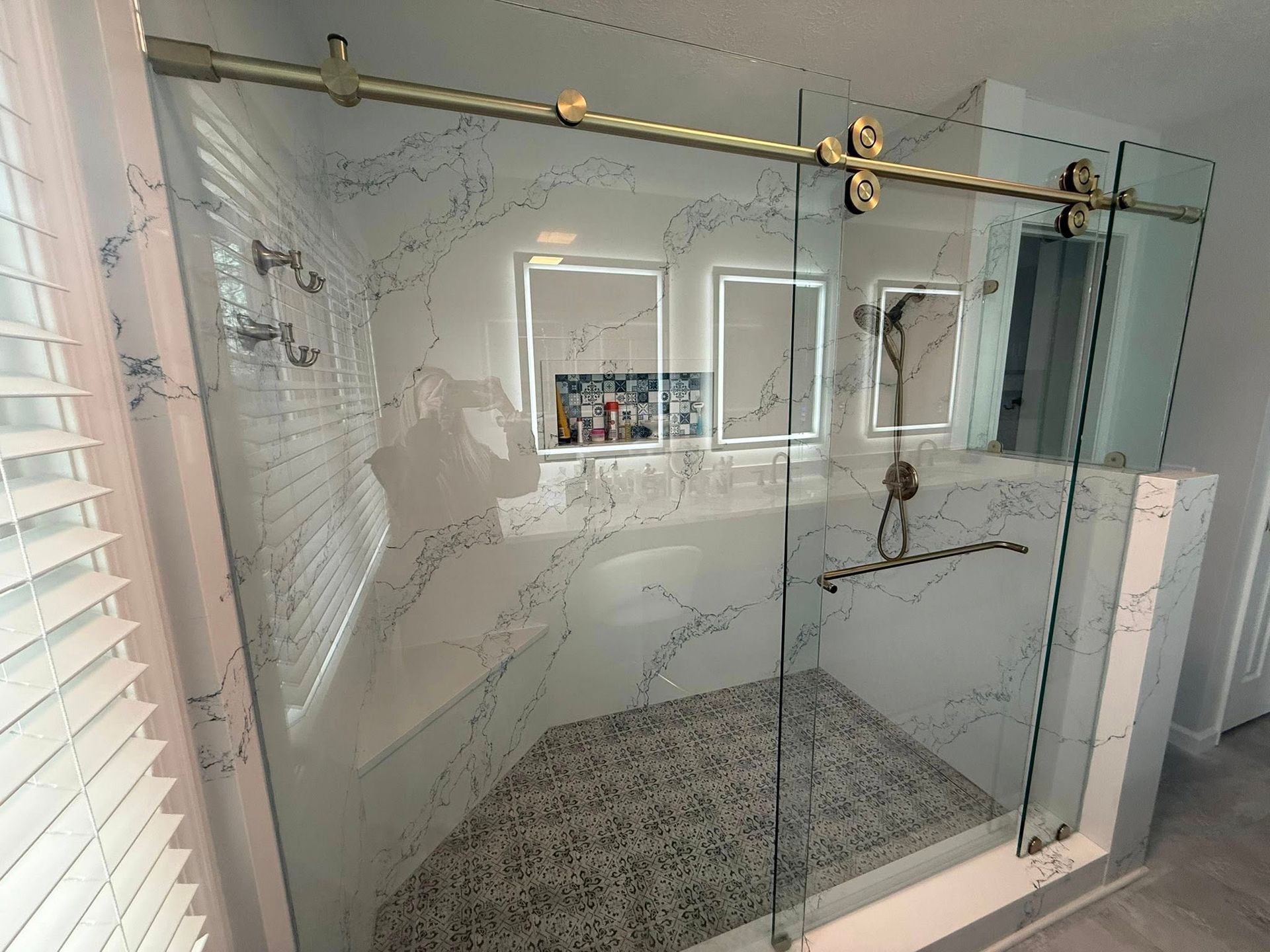Glass-enclosed shower with brass rail, patterned tile floor, and white tiled walls with windows and built-in shelves
