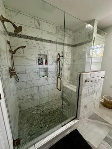 Glass shower with white marble tile, bronze fixtures, built-in shelf, and black floor mat