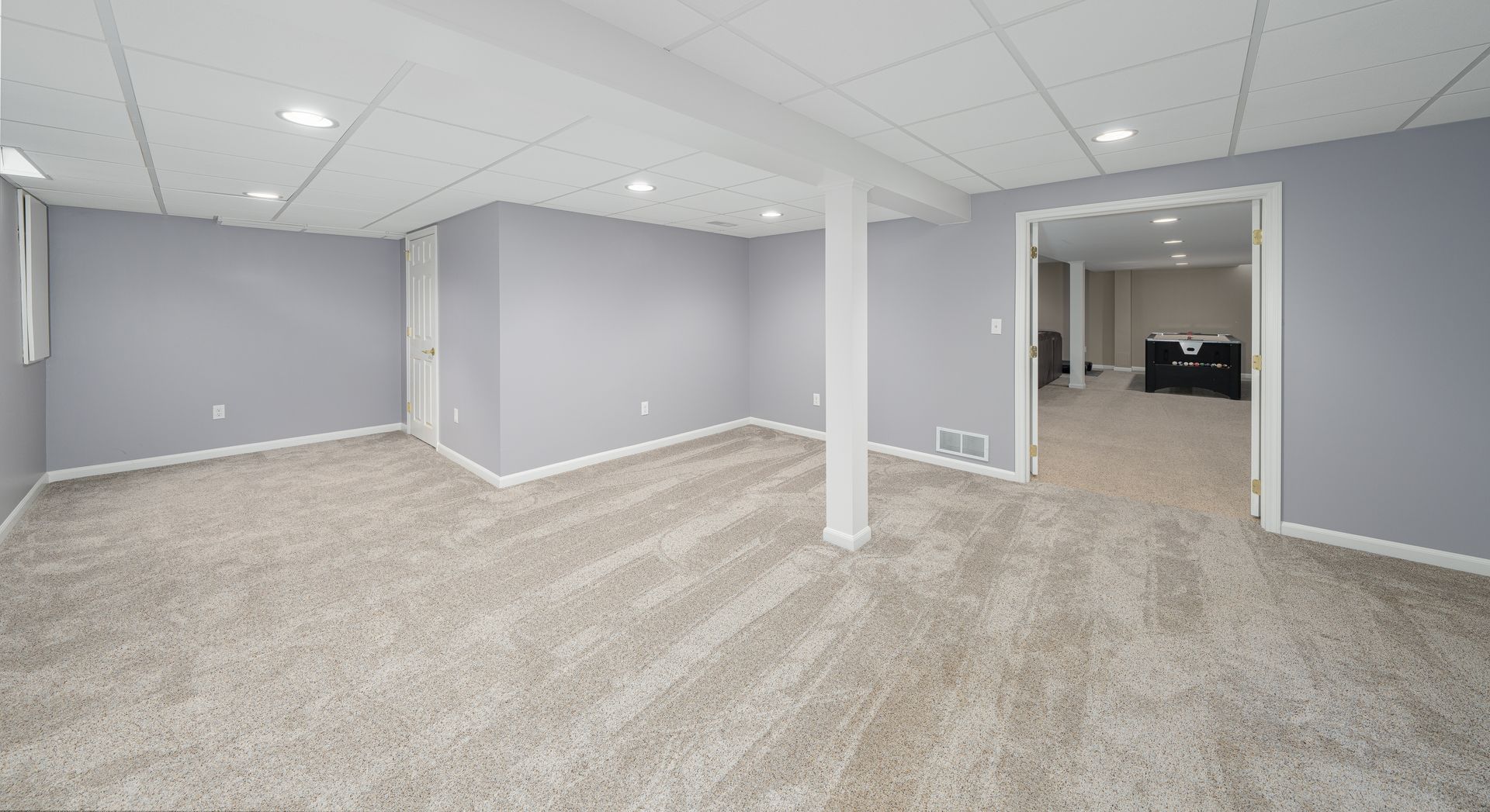 An unfurnished basement room with light gray walls, a tan carpet, and white drop ceilings leading into an adjacent room.