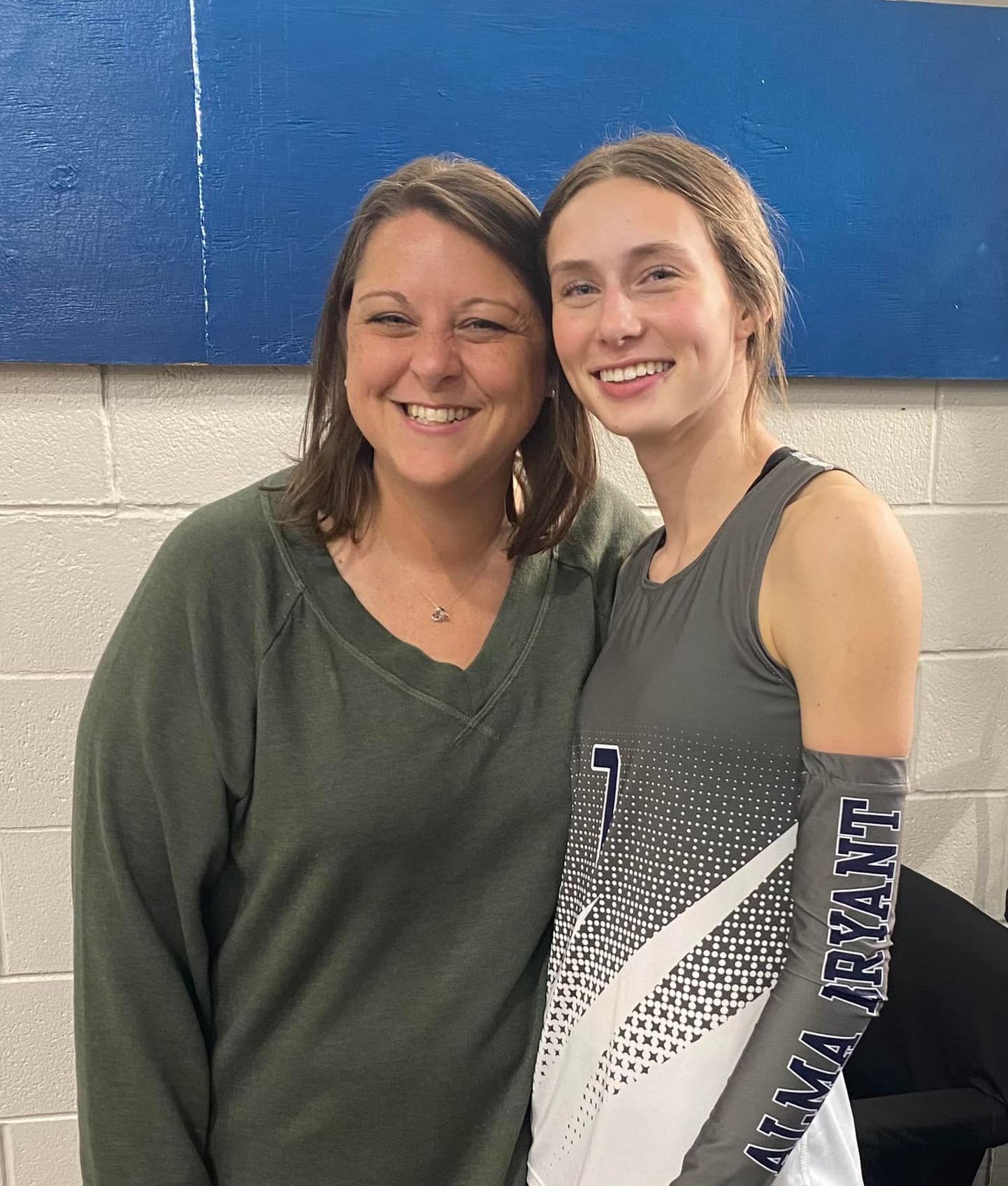 Woman with another young woman, smiles, posing against a blue wall. 