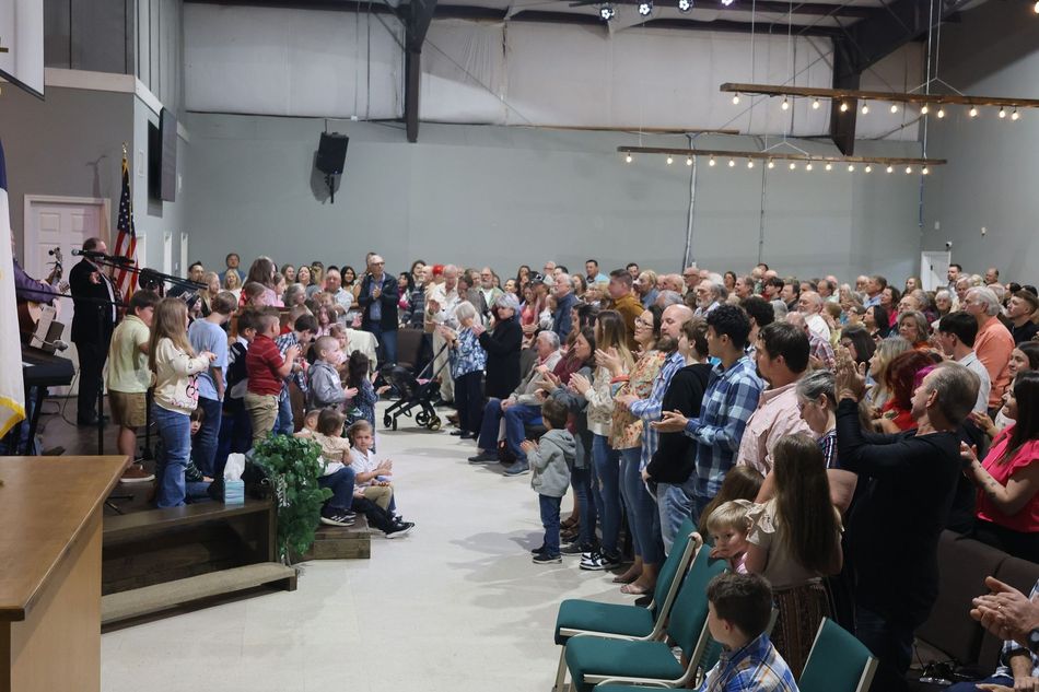 Large crowd applauding in a brightly lit room. A few people stand near a podium.