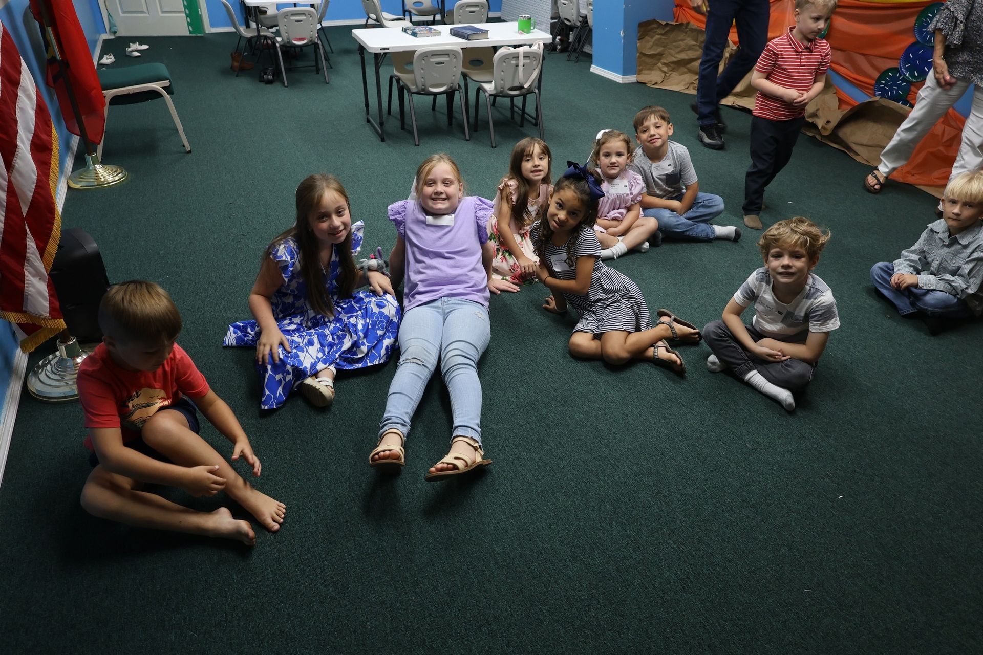 Children gathered on carpeted floor in a room. Some sit, some stand, and one runs.